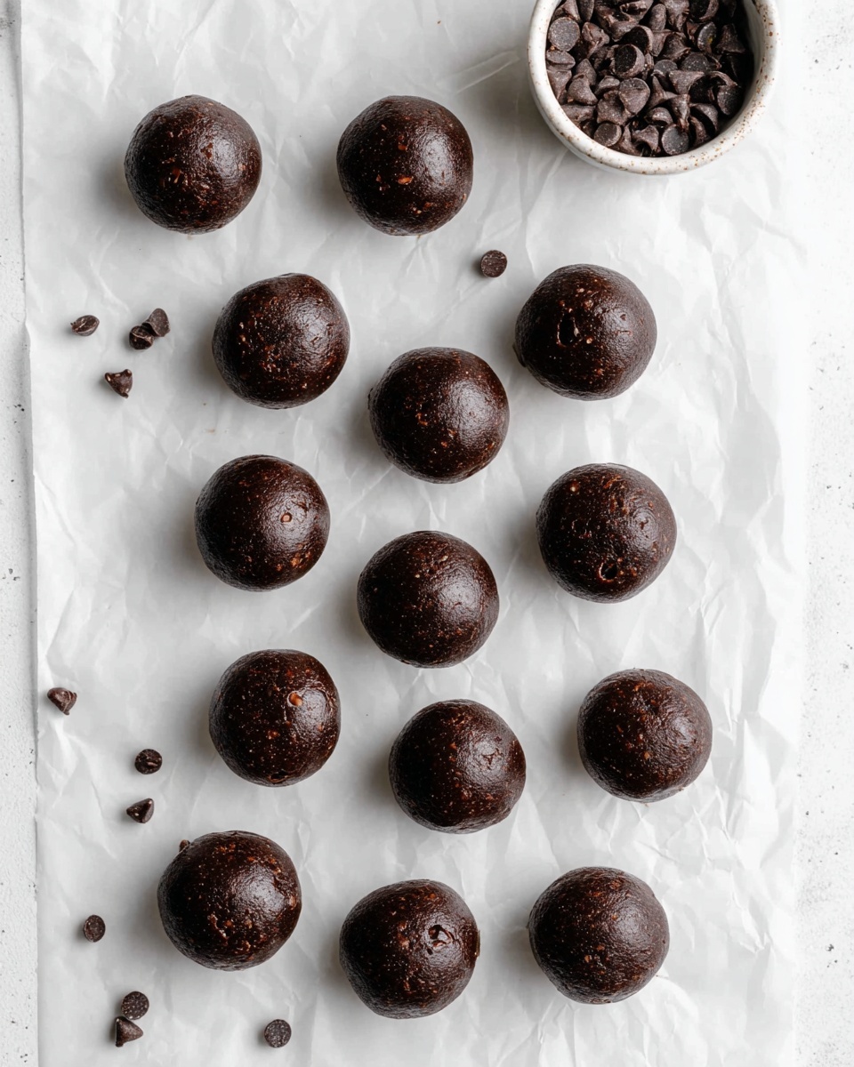 The image shows about fifteen round dark chocolate balls evenly placed on a slightly crumpled white paper over a white marbled surface. The balls have a smooth, glossy texture with a few small chocolate chips embedded on their surface, adding tiny bits of texture and lighter brown spots. To the top right corner, there is a white bowl filled with small dark chocolate chips, some of which are scattered around the balls on the white paper. The overall look is clean and simple with a natural lighting that highlights the shine on the balls. Photo taken with an iphone --ar 4:5 --v 7