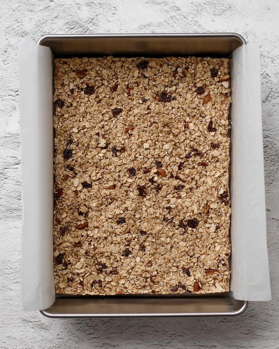 The image shows a rectangular metal baking pan lined with white parchment paper filled with an even layer of a grainy oat mixture. The mixture is light brown with visible pieces of darker nuts or dried fruits spread throughout, giving it a rough textured look. The pan is placed on a white marbled surface, adding contrast to the pan's metal edges and the filled oat layer inside. photo taken with an iphone --ar 4:5 --v 7