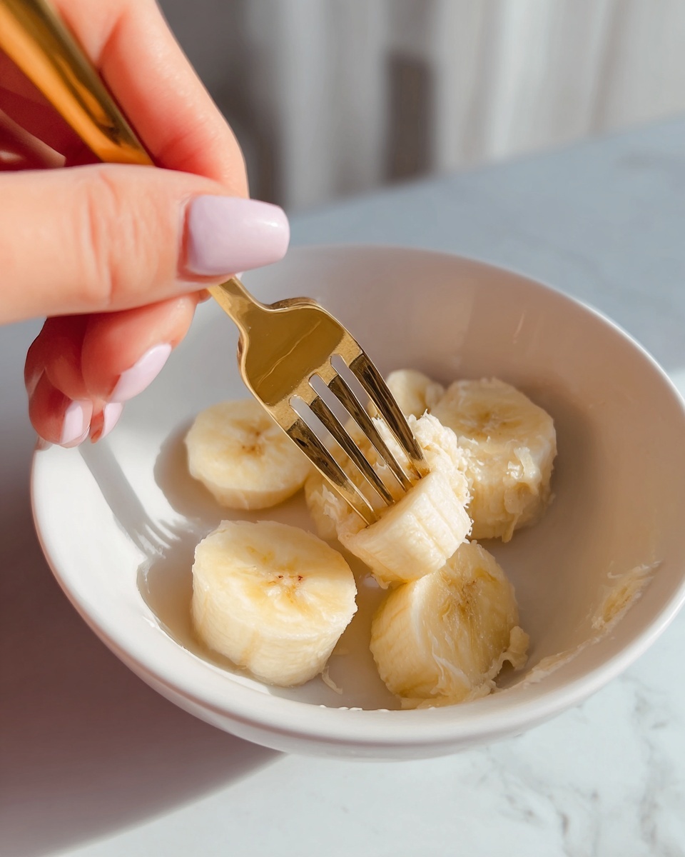 A close-up image shows a white bowl on a white marbled surface, with three pieces of peeled banana inside. A woman's hand with light pink nail polish holds a gold fork mashing one banana piece in the bowl. The bananas look soft and creamy with a pale yellow color. The background is softly blurred, emphasizing the action in the bowl. photo taken with an iphone --ar 4:5 --v 7