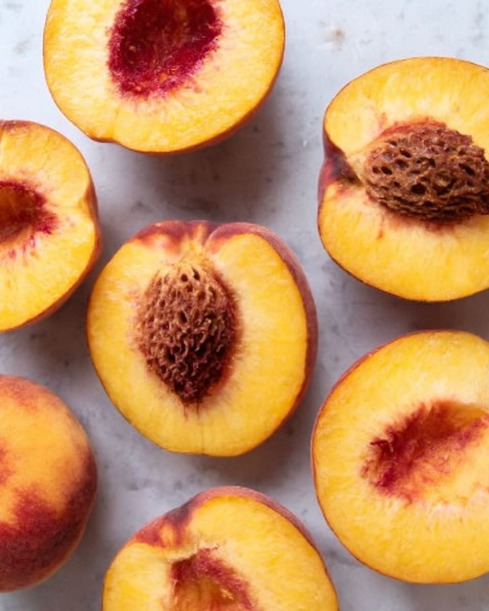The image shows several peach halves placed on a white marbled surface. Each peach is golden-yellow with a red blush near the center, exposing the brown textured pit in the middle. The skin edges are reddish-orange with a smooth texture, while the inside flesh looks juicy and soft. The peaches are arranged in a casual, scattered manner, showing a natural and fresh appearance. Photo taken with an iphone --ar 4:5 --v 7
