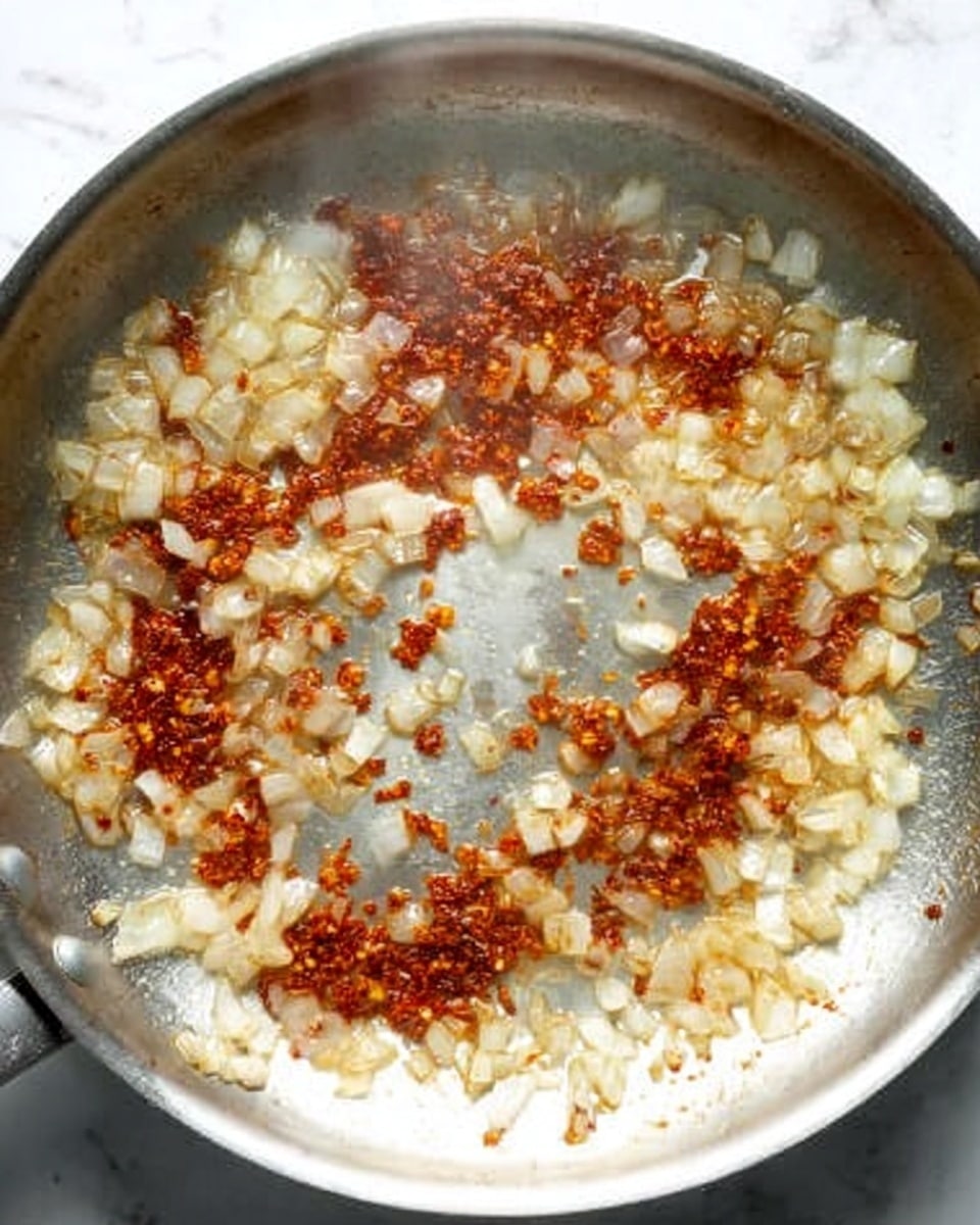 The image shows a silver frying pan on a white marbled surface, with cooked diced onions spread mostly in the lower half and some reddish-brown cooked spices or sauce pieces scattered more densely along the upper half. The inside of the pan has a slightly shiny texture with small bits stuck due to cooking, creating a mix of white, light golden, and reddish-brown colors, giving a clear view of the pan’s metallic surface in the center. The steam rising lightly suggests the food is hot and freshly cooked. Photo taken with an iphone --ar 4:5 --v 7