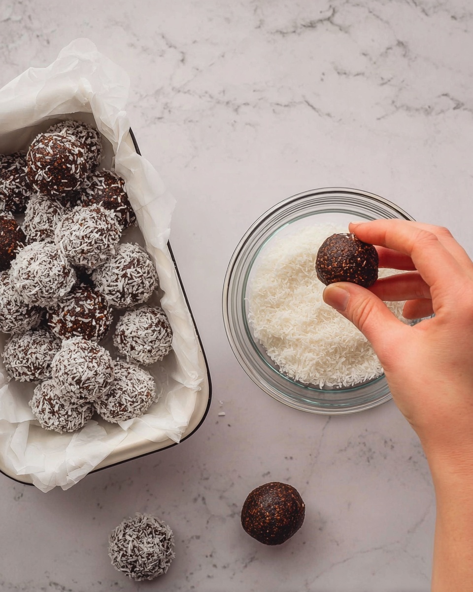A woman's hand is holding a dark brown round ball with a rough texture and dipping it into a small clear glass bowl filled with white shredded coconut flakes. To the left, there is a white container lined with white parchment paper holding several more of the dark brown balls, some of which are partially covered in the white coconut flakes, creating a contrast between the rough dark brown and the soft white texture. The scene is set on a white marbled surface. Photo taken with an iphone --ar 4:5 --v 7