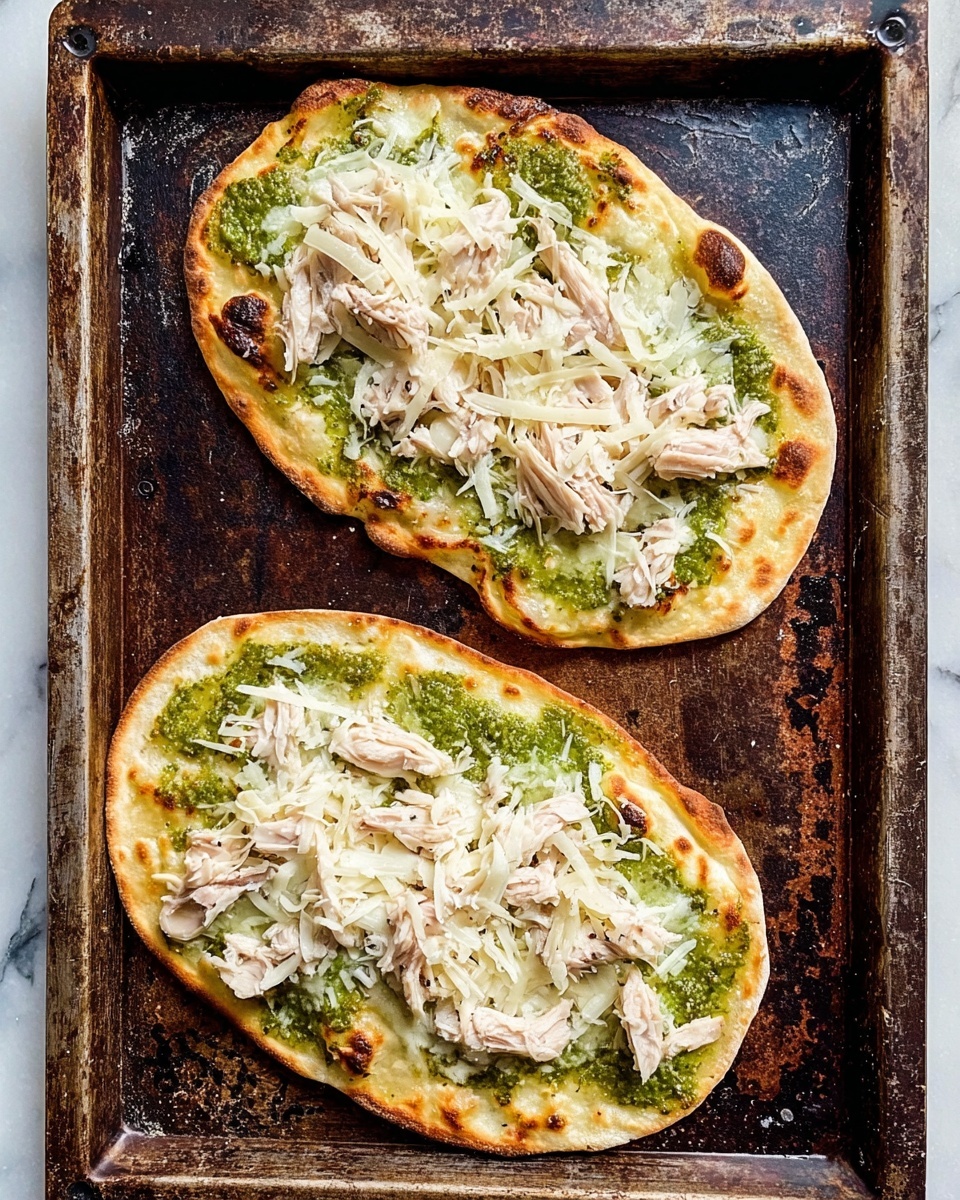 The image shows two oval flatbreads on a dark, worn baking tray. Each flatbread has three visible layers: the bottom layer is a light golden, slightly puffed flatbread with darker toasted spots around the edges; the middle layer is a spread of green sauce covering most of the flatbread surface with a textured, slightly chunky look; the top layer is shredded white cheese sprinkled evenly across the flatbread with small piles of light pink shredded chicken placed randomly over the cheese. The baking tray sits on a white marbled surface. photo taken with an iphone --ar 4:5 --v 7