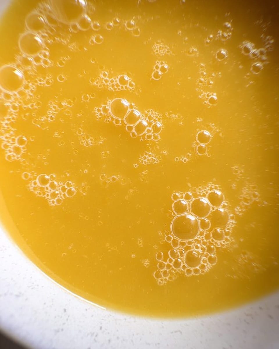 A close-up view of a smooth, thick yellow soup filling a white bowl, with small bubbles scattered on the surface, placed on a white marbled texture background photo taken with an iphone --ar 4:5 --v 7