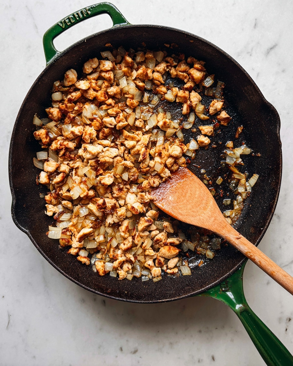 A black cast iron pan with a green handle is shown on top of a white marbled surface. Inside the pan, there is a mix of small, browned pieces of cooked chicken and diced onions, all lightly browned and mixed together. A wooden spoon with a smooth texture is resting inside the pan, partially stirring the ingredients. The pan shows some areas of cooked bits sticking lightly to the bottom edges. The scene captures a warm, homey cooking moment. photo taken with an iphone --ar 4:5 --v 7