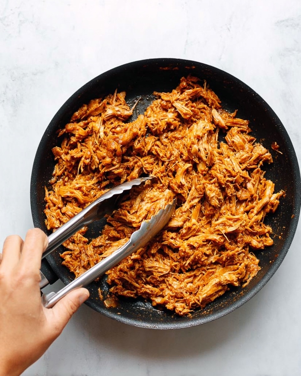 The image shows a black frying pan filled with shredded, cooked meat that is dark orange-brown in color and has a slightly moist texture, with small pieces clumped together. The pan is held by a woman's hand using metal tongs to lift some of the meat, positioned on the lower left side of the pan. The background surface is a white marbled texture. Photo taken with an iphone --ar 4:5 --v 7