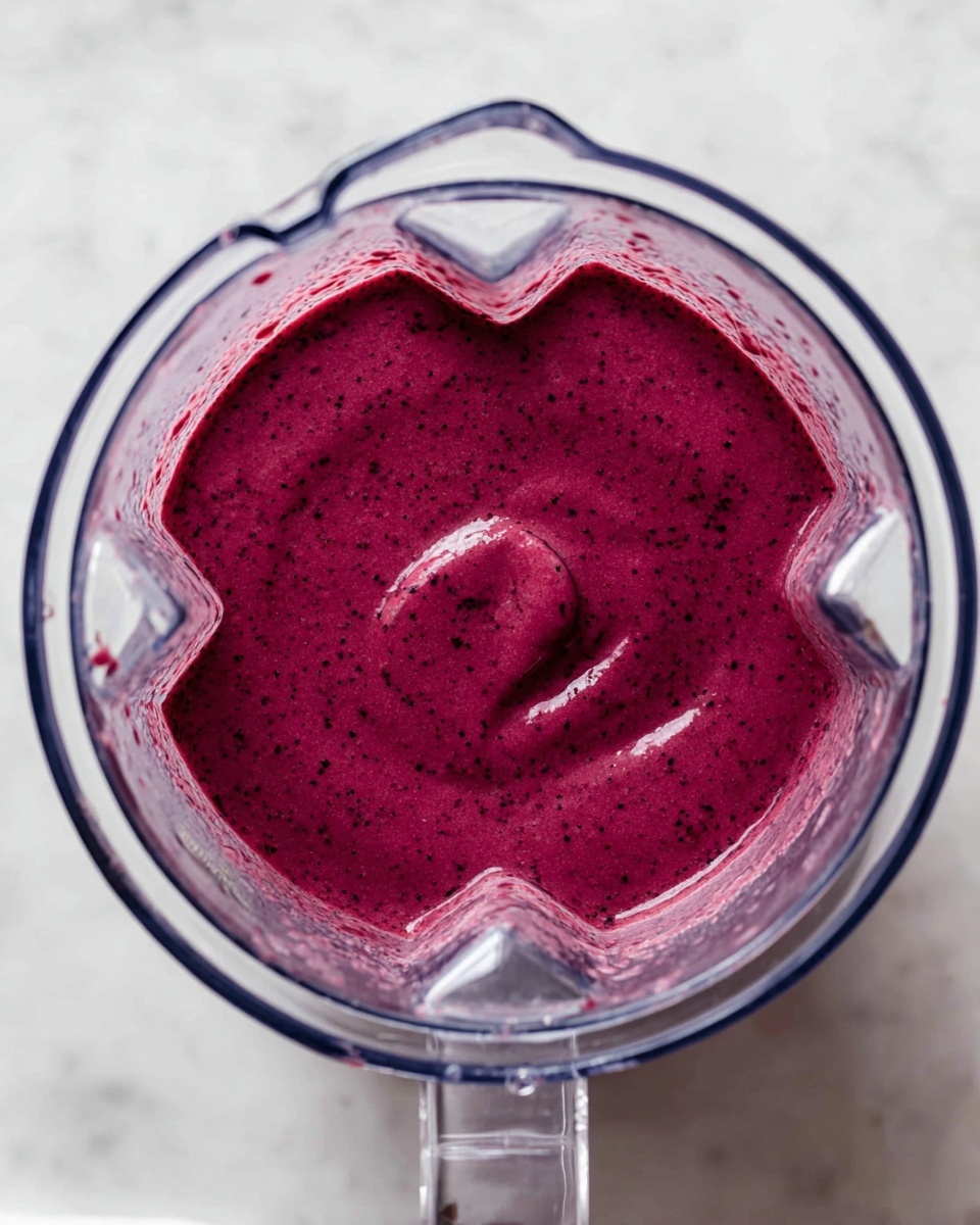 A clear blender container filled with a smooth, thick, deep magenta liquid with tiny darker specks scattered throughout. The blender is set on a white marbled surface, and the liquid reaches nearly to the top but leaves a small empty space. The blender lid is partially visible on the right side of the image, also clear plastic. The photo is taken from directly above, showing the even texture and vibrant color of the blended mixture photo taken with an iphone --ar 4:5 --v 7