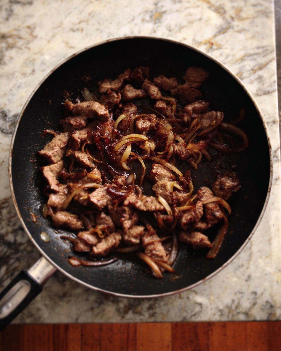 The image shows a black pan on a white marbled surface, filled with cooked sliced meat. The meat pieces are brown with some darker edges, looking tender and slightly juicy. There are small bits of thin, light brown onion mixed throughout. The pan handle is visible, and the whole scene has warm lighting. Photo taken with an iphone --ar 4:5 --v 7