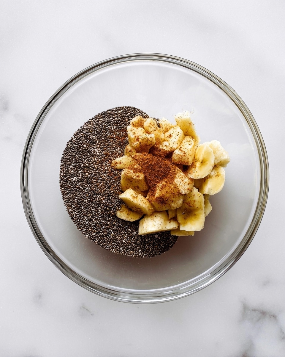 A clear glass bowl holds three layers of ingredients on a white marbled surface: the bottom layer is a thick pile of small, round, dark chia seeds positioned on the left side; the top right side shows a cluster of yellowish chopped banana pieces sprinkled with light brown cinnamon powder; the layers are evenly placed next to each other without mixing, and the bowl edges are clean and clear, showing the ingredients inside well, photo taken with an iphone --ar 4:5 --v 7