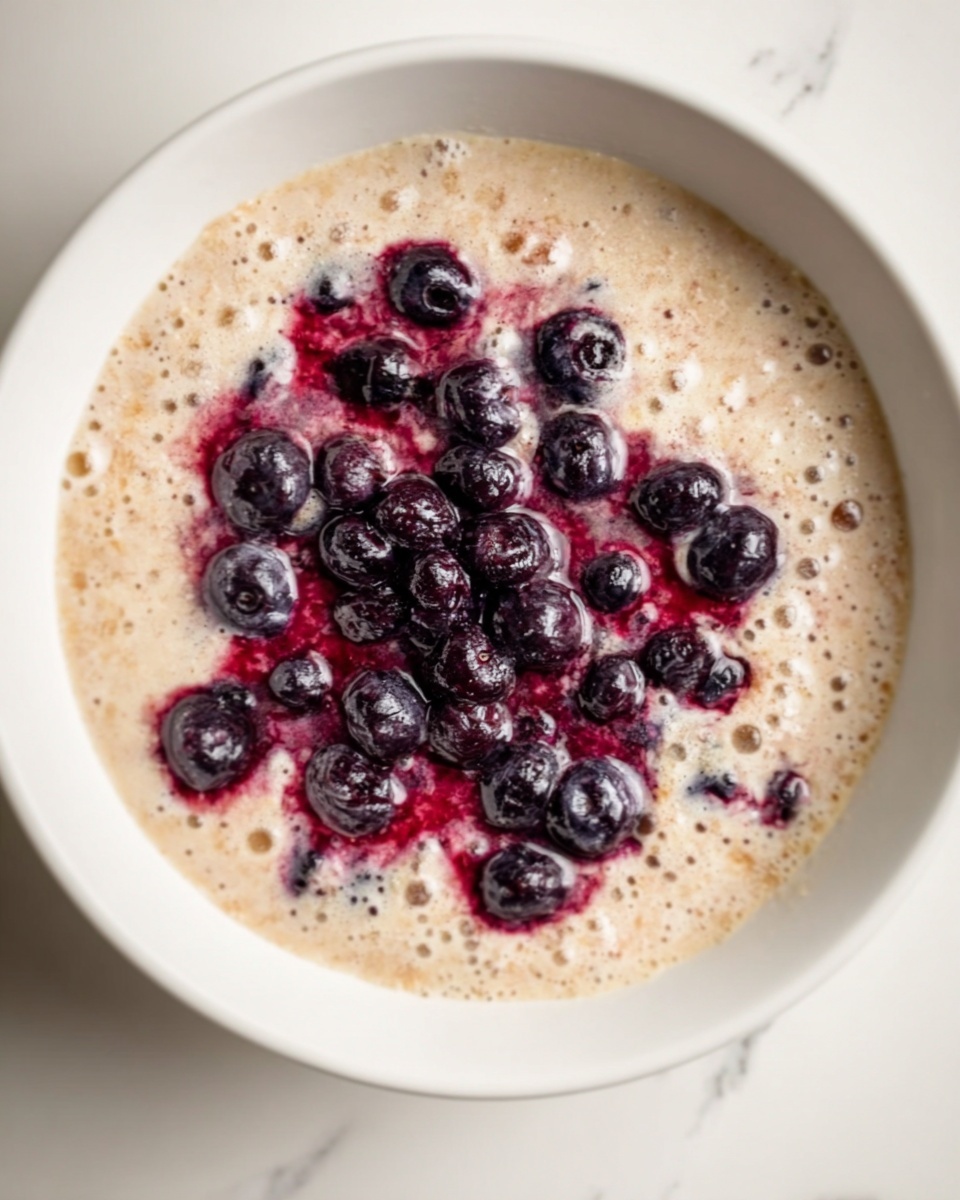 A white bowl filled with a thick pancake batter that has a light beige color and small bubbles on the surface. On top, there are several clusters of dark purple blueberries, some bursting and spreading a bit of deep red juice over the batter, creating a slight contrast. The bowl rests on a white marbled surface. photo taken with an iphone --ar 4:5 --v 7