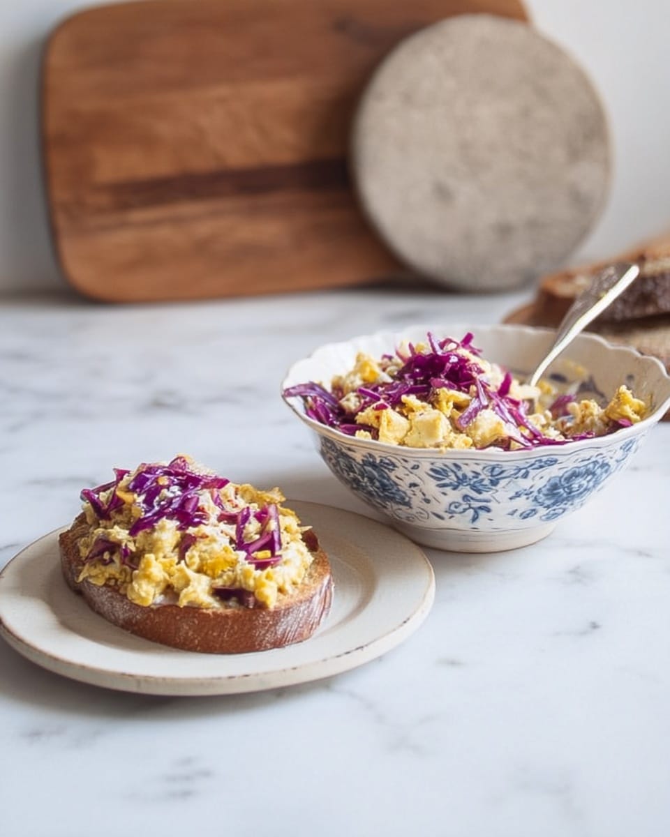 The image shows a small piece of toast placed on a white plate, topped with a colorful mix of yellow scrambled eggs and purple cabbage or onions, creating a layered look with the eggs on the bottom and the purple vegetables on top. Next to it, there is a decorative white and blue bowl filled with the same egg and purple vegetable mixture, showing a combination of soft, scrambled eggs and thin, shredded purple pieces. A spoon rests inside the bowl. The background consists of a clean white marbled surface with a wooden cutting board and a stone round in the back. The lighting is soft and natural. Photo taken with an iphone --ar 4:5 --v 7