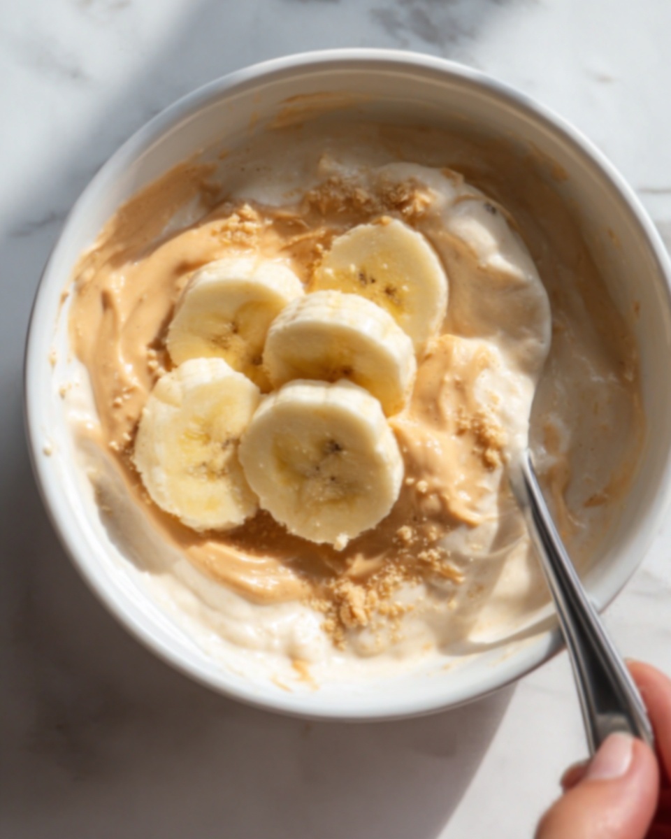 A close-up view of a white bowl filled with creamy, light beige yogurt mixed with smooth peanut butter that creates a slightly thick texture. On top, several slices of yellow banana are placed neatly, half-buried in the yogurt mix, showing their soft, smooth surface. A silver spoon is partially inside the bowl, held by a woman's hand, blending into the creamy mixture. The bowl rests on a white marbled surface with soft natural lighting highlighting the slight gloss of the yogurt. Photo taken with an iphone --ar 4:5 --v 7