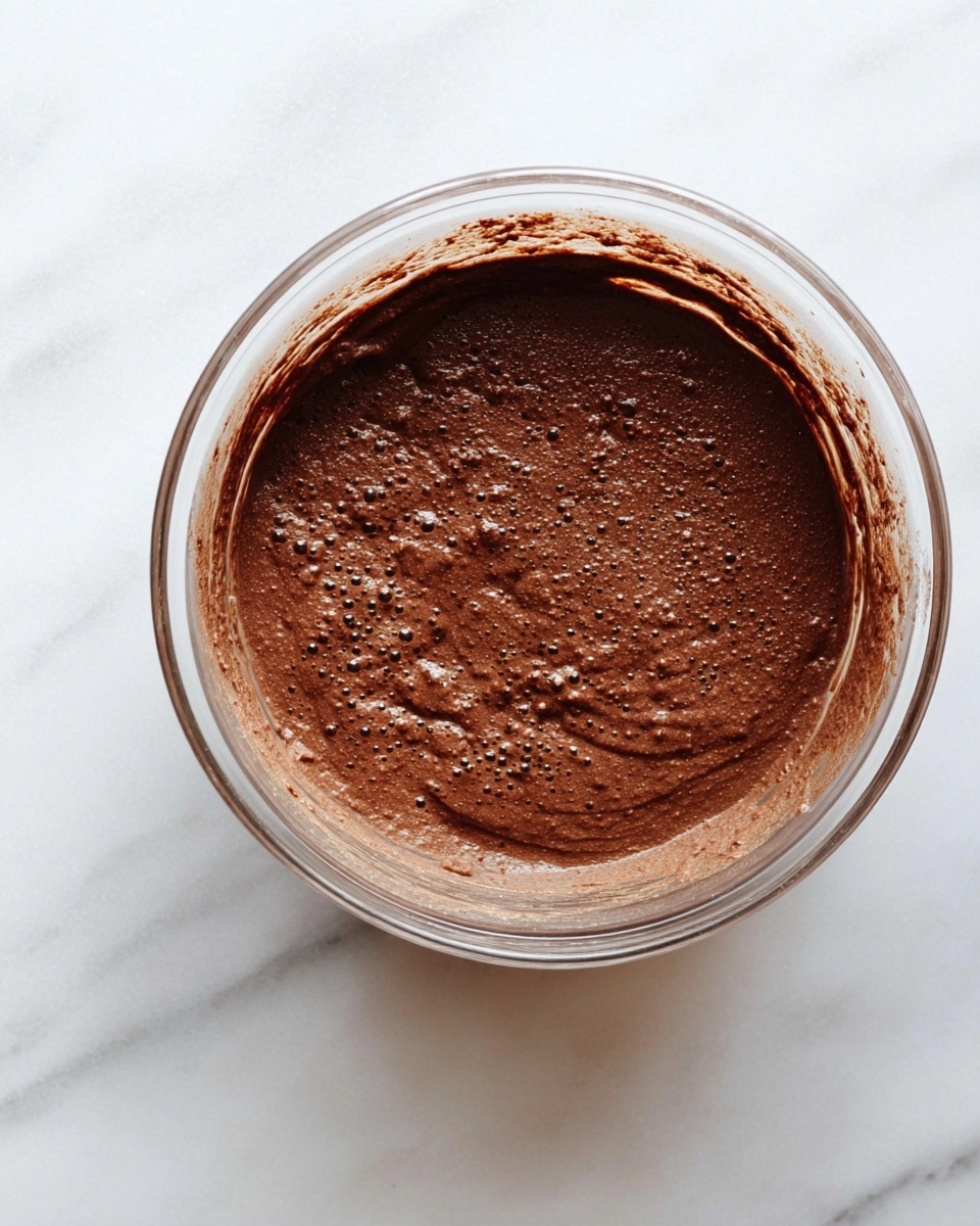 A clear glass bowl filled with a smooth, thick chocolate mixture that has a slightly bubbly texture on the surface. The chocolate batter is dark brown, evenly spread inside the bowl with some splashes on the inner sides. The bowl is placed on a white marbled surface, with soft natural lighting highlighting the rich color and texture of the chocolate mix. photo taken with an iphone --ar 4:5 --v 7