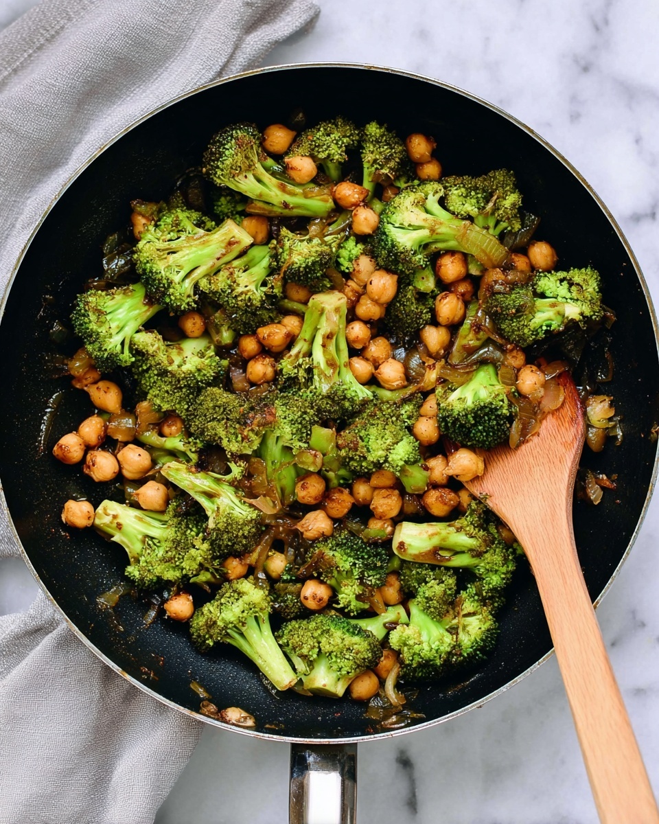 A black frying pan filled with cooked broccoli and chickpeas is shown from above. The broccoli is bright green with some browned spots, and the chickpeas are golden brown and scattered evenly throughout. There are visible bits of cooked onion mixed in, adding texture and a slight shine from the cooking oil. A wooden spatula rests in the pan on the right side. The pan sits on a white marbled surface with a light grey kitchen cloth partially visible in the top left corner. Photo taken with an iphone --ar 4:5 --v 7