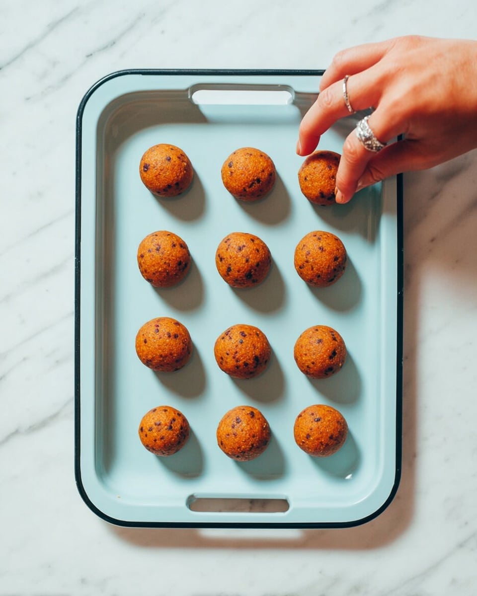 Fifteen small, round orange-brown balls with dark specks are neatly placed in a 3 by 5 grid on a light blue tray with black edges and two small handles. A woman's hand with a silver ring is gently picking up one ball from the upper left corner. The tray sits on a white marbled surface. The lighting is soft and natural, casting faint shadows beneath each ball. photo taken with an iphone --ar 4:5 --v 7