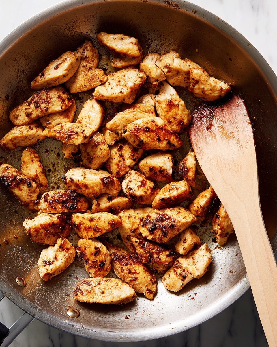 A close-up top view of a shiny silver pan containing small, uneven pieces of cooked chicken in different shades of golden brown with dark grilled spots. The chicken pieces are spread mostly on the left half of the pan, with some pieces flicked towards the bottom center of the pan. A smooth wooden spatula with light brown color rests on the right side of the pan, touching the chicken. The background is a white marbled texture. photo taken with an iphone --ar 4:5 --v 7
