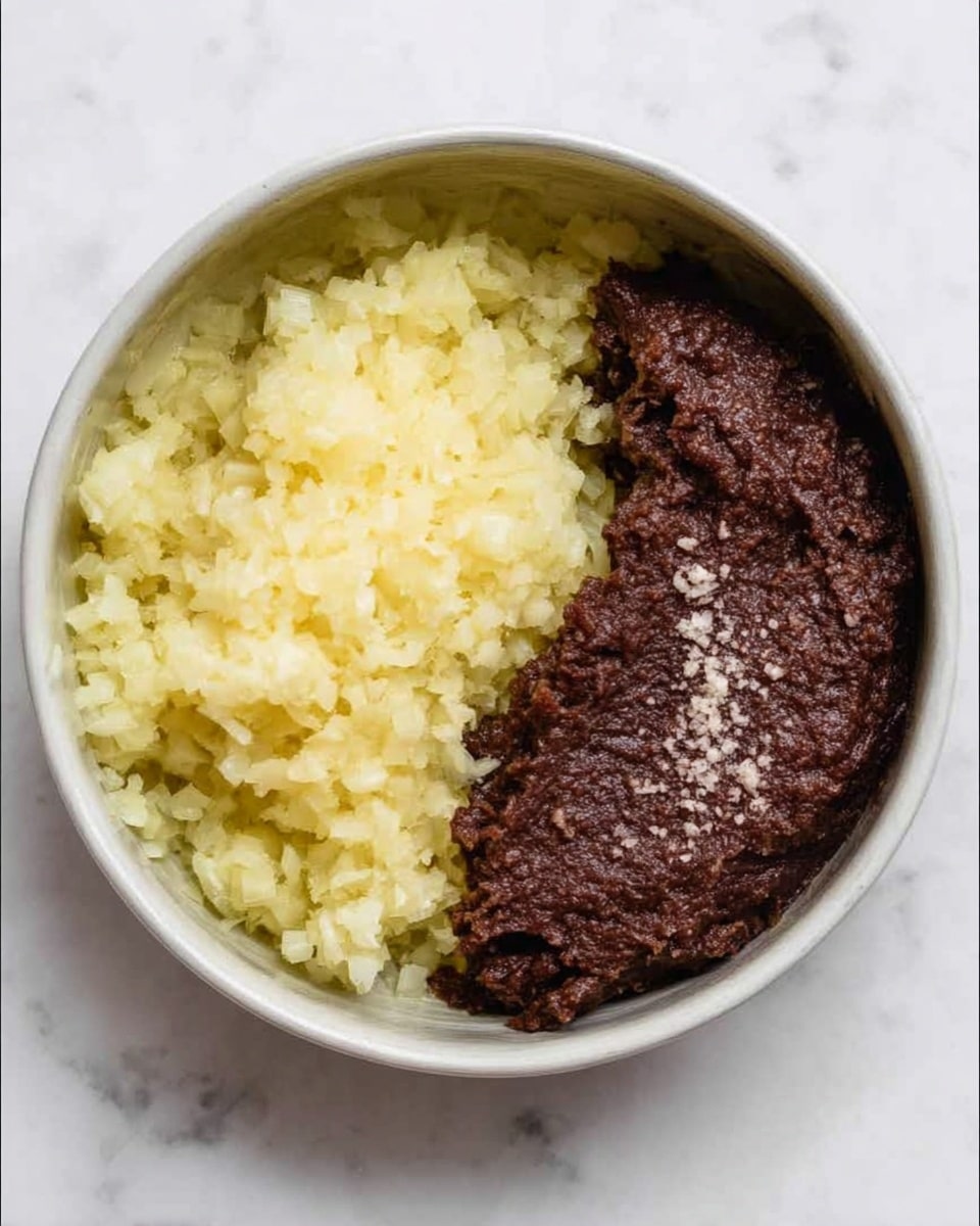 A white bowl with two main parts inside: on the left, there is a pile of finely chopped light yellow onions with a soft texture, and on the right, there is a thick dark brown paste with a slightly rough surface and some small white granules sprinkled on top. The two parts sit side by side, almost filling the bowl, on a white marbled surface. Photo taken with an iphone --ar 4:5 --v 7