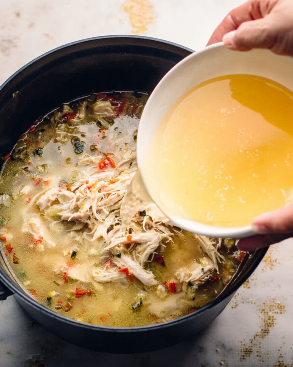 A close-up view of a large black cooking pot filled with shredded pale chicken pieces and small bits of vegetables in light broth. A white bowl is tilted above the pot, pouring a light yellow liquid with visible bits of red and green into the pot. A woman's hand is holding the white bowl, and the pot is sitting on a white marbled surface with small specks of liquid around it. The scene shows the moment of adding the liquid to the cooking pot. photo taken with an iphone --ar 4:5 --v 7