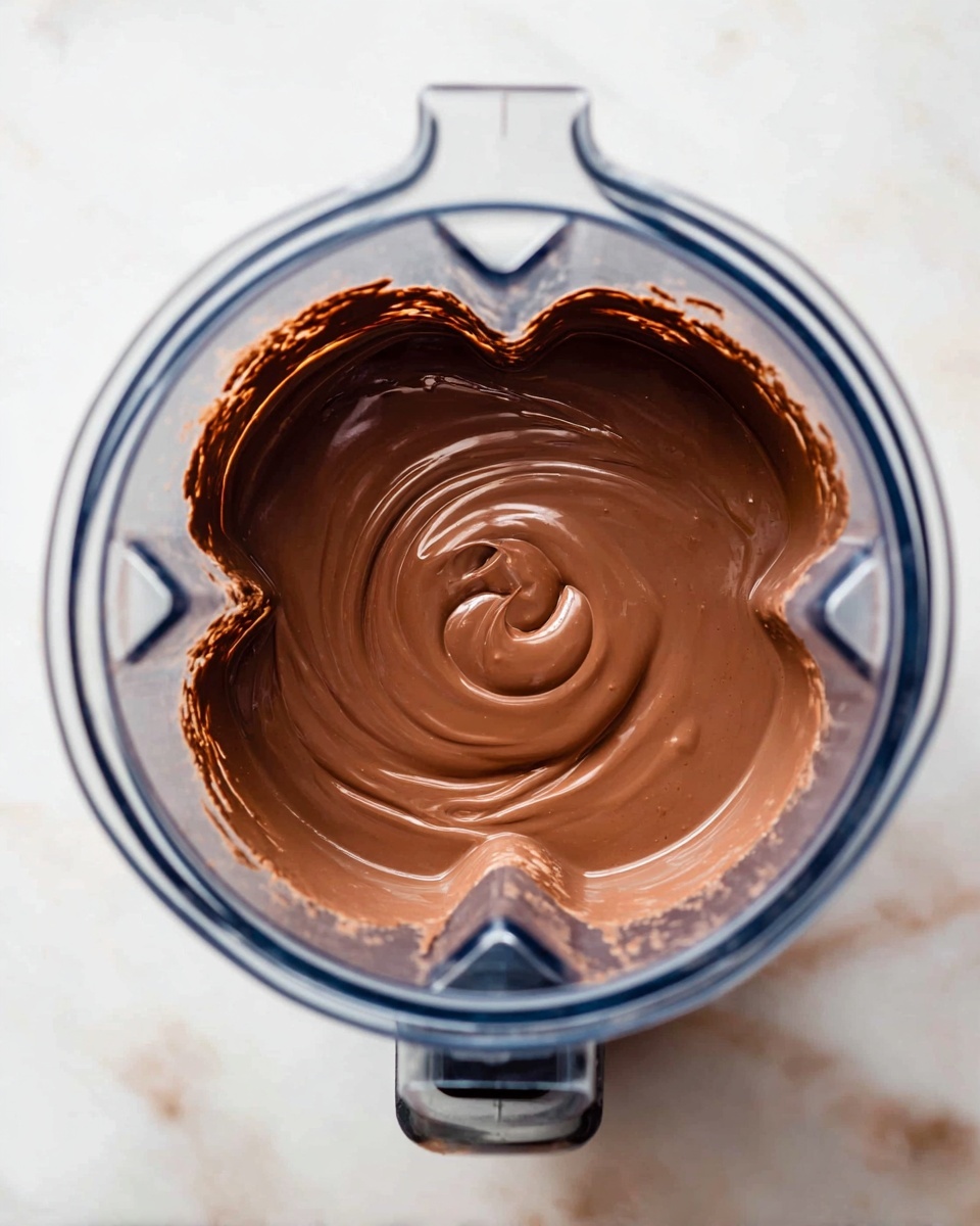 A top view of a clear blender jar filled with smooth, shiny chocolate mixture swirling in the center. The chocolate has a thick, creamy texture with a rich brown color. The blender's blades are visible beneath the surface, and the jar sits on a white marbled surface. photo taken with an iphone --ar 4:5 --v 7