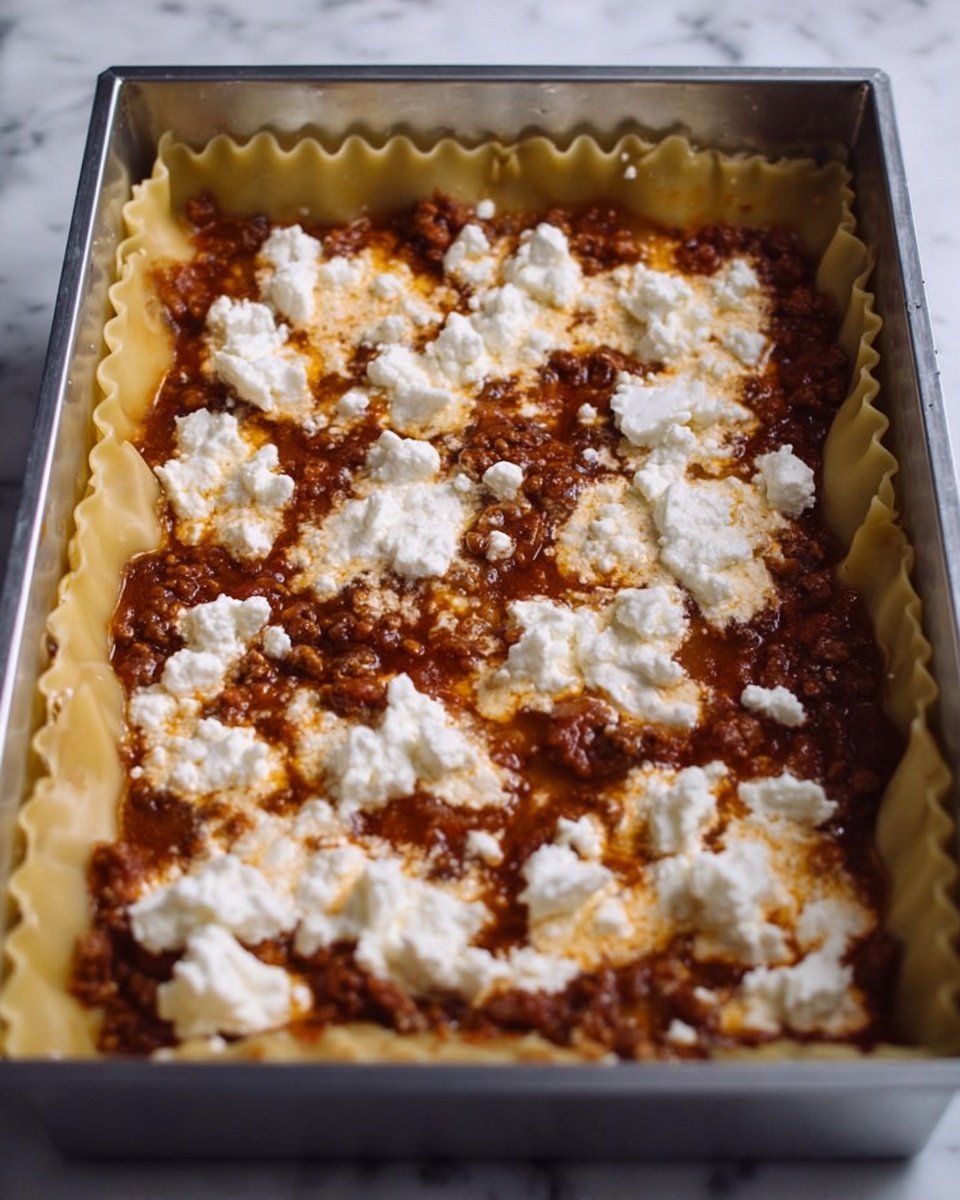 The image shows a close-up of a metal baking dish with a layer of lasagna inside. The bottom layer is pasta sheets, with a brown meat and tomato sauce spread evenly on top. Over the sauce, there are scattered small dollops of white ricotta cheese. The pasta edges are folded slightly upward over the sauce. The background is a white marbled texture. photo taken with an iphone --ar 4:5 --v 7