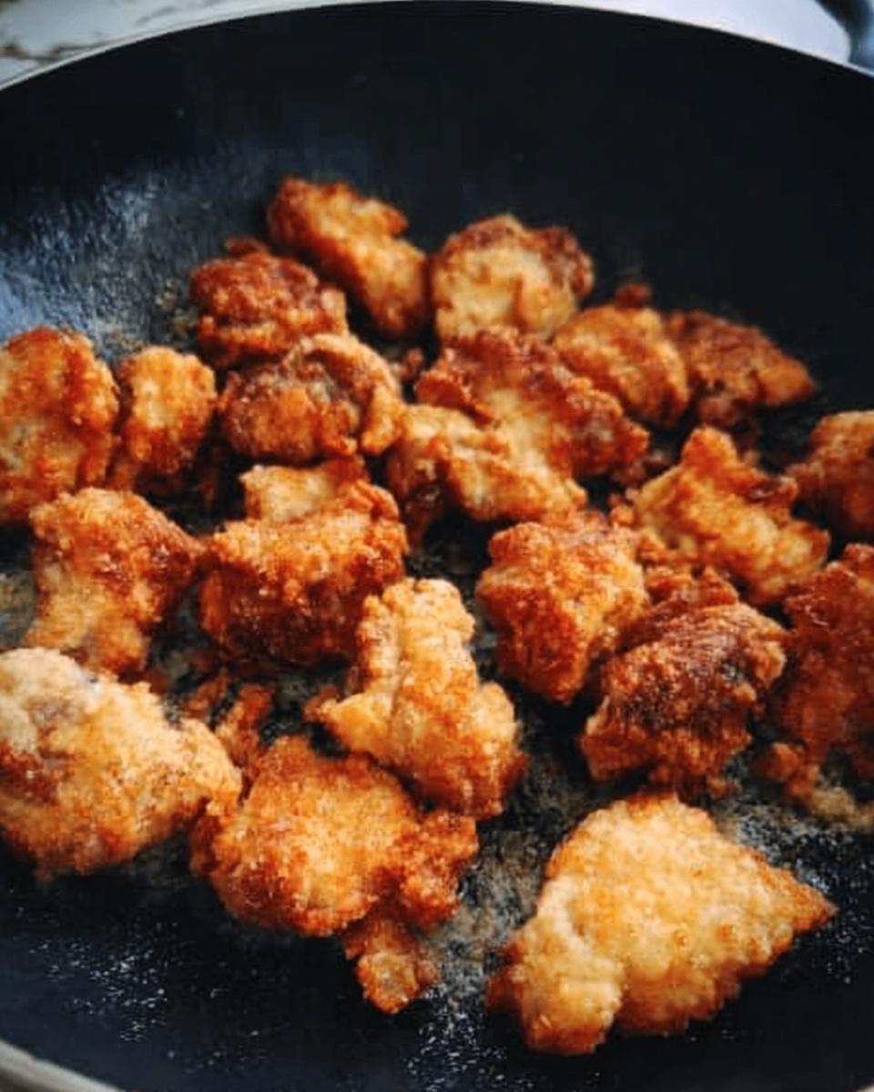 The image shows small pieces of golden brown fried chicken in a black pan with a slightly rough texture. The chicken pieces are irregular in shape, with a crunchy, crispy surface, some parts darker and some lighter. The pan fills the frame, and the background is a white marbled texture. Photo taken with an iphone --ar 4:5 --v 7