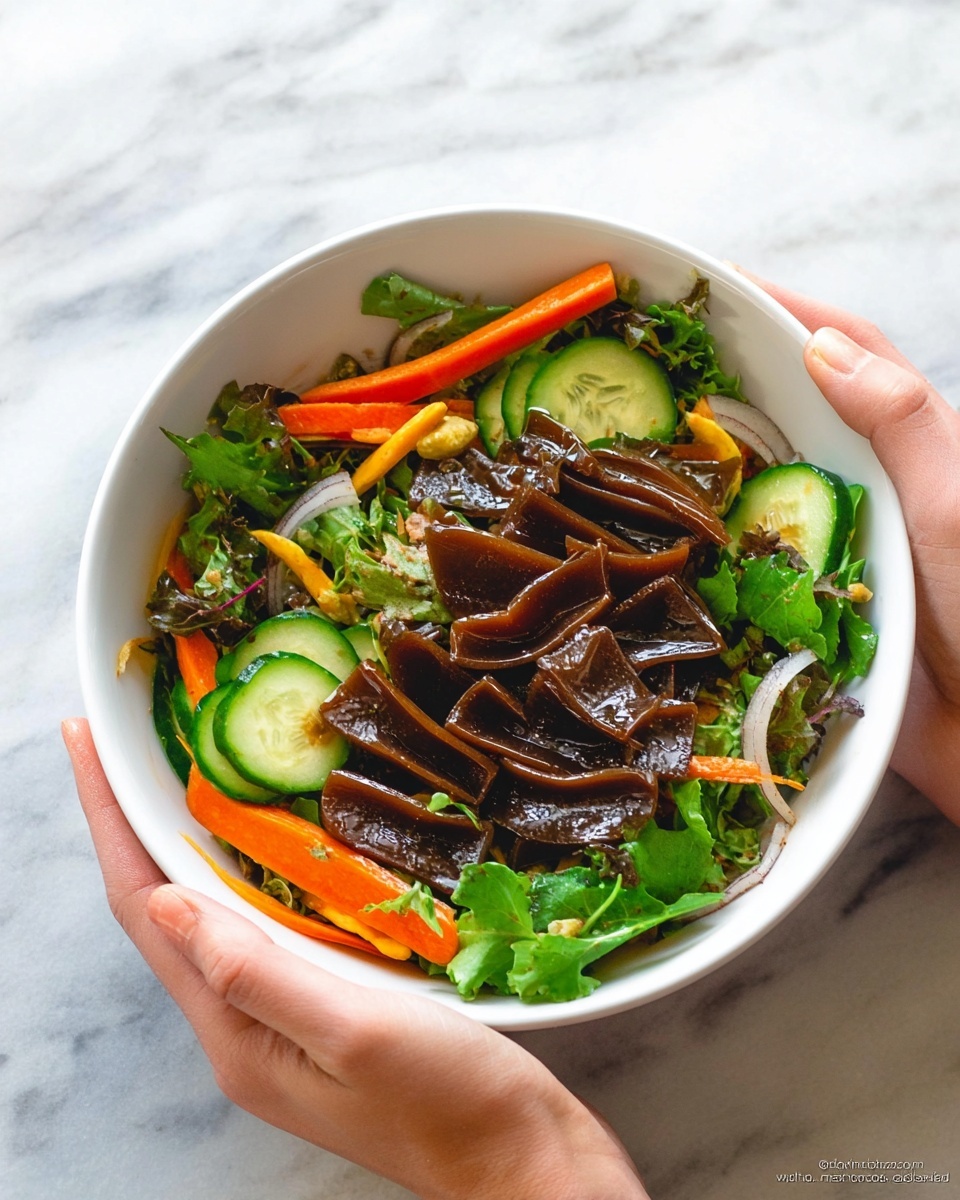 A white bowl held by a woman's hands contains a colorful salad with several layers: the top layer has dark brown, wavy rectangular jelly-like pieces spread evenly; underneath are bright green cucumber slices cut into halves and slender orange carrot sticks scattered throughout; mixed in with these are light green leafy vegetables and thin slices of white onion with a hint of translucence. All ingredients look fresh and slightly glossy, showing a light dressing. The bowl is shown against a white marbled surface. Photo taken with an iphone --ar 4:5 --v 7