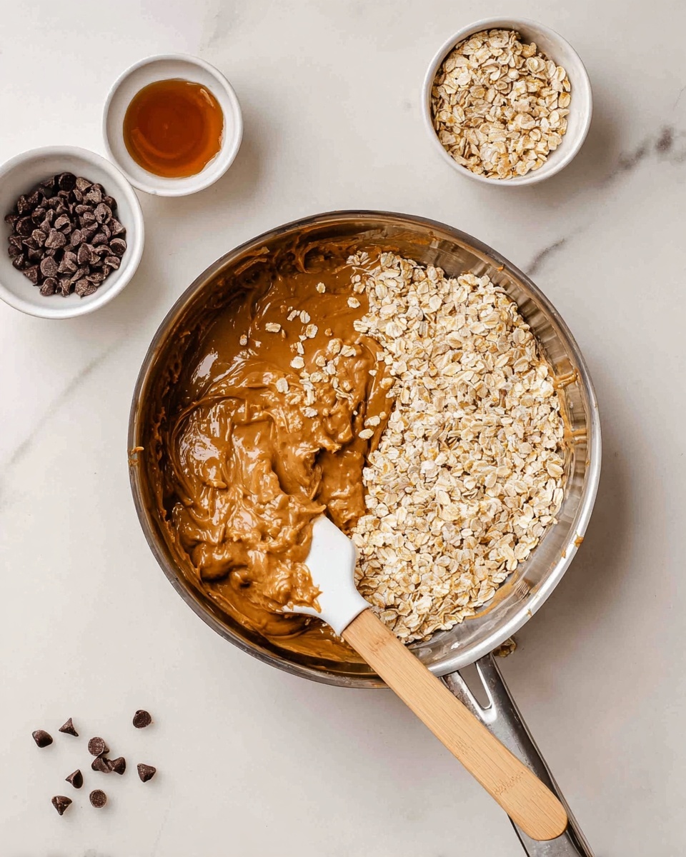 A shiny metal pan holds a mixture of light brown peanut butter and raw oats. The peanut butter layer is smooth and thick, sitting mostly on the left side of the pan, while dry oats are sprinkled over the right side and partially mixed in. A white and wooden spatula rests inside the pan on the right, coated with the sticky peanut butter and some oats. Around the pan on a white marbled surface, there are three small white bowls containing light rolled oats, dark chocolate chips, and a small amount of liquid syrup. A few chocolate chips are scattered loosely near the bowls. photo taken with an iphone --ar 4:5 --v 7