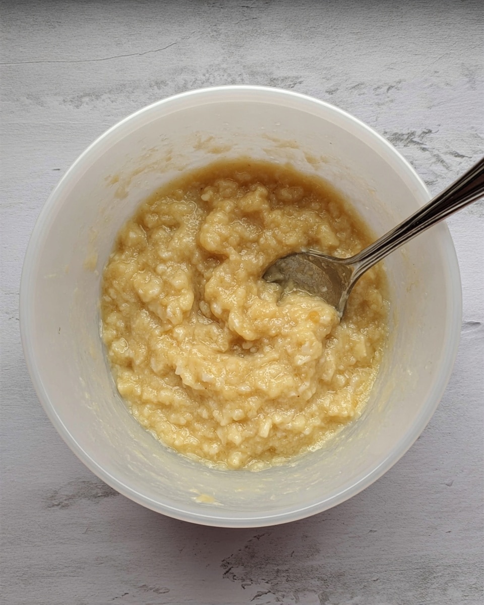 A top view of a white translucent bowl showing a single layer of mashed banana mixture with a chunky and soft texture, yellowish-beige in color, with small bits visible throughout; a silver spoon is partially submerged in the mixture on the right side of the bowl, all placed on a white marbled surface. photo taken with an iphone --ar 4:5 --v 7