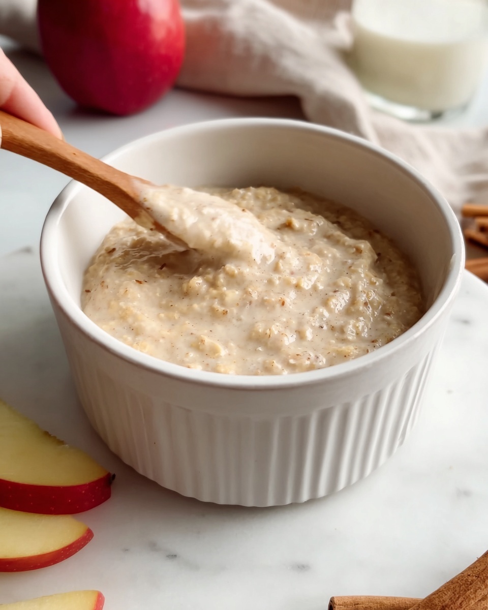 A thick creamy oatmeal mixture with small chunks is inside a round white ceramic bowl with ridges on the outside. The oatmeal has a light beige color with tiny bits visible, showing texture. The bowl is placed on a white marbled surface, and pieces of red and yellow apple slices sit nearby, adding color contrast. Part of a cinnamon stick is visible on the side. A blurred woman's hand is holding a wooden spoon above the bowl, stirring the oatmeal gently. In the background, there is a red apple and a glass of white liquid, softly focused. photo taken with an iphone --ar 4:5 --v 7