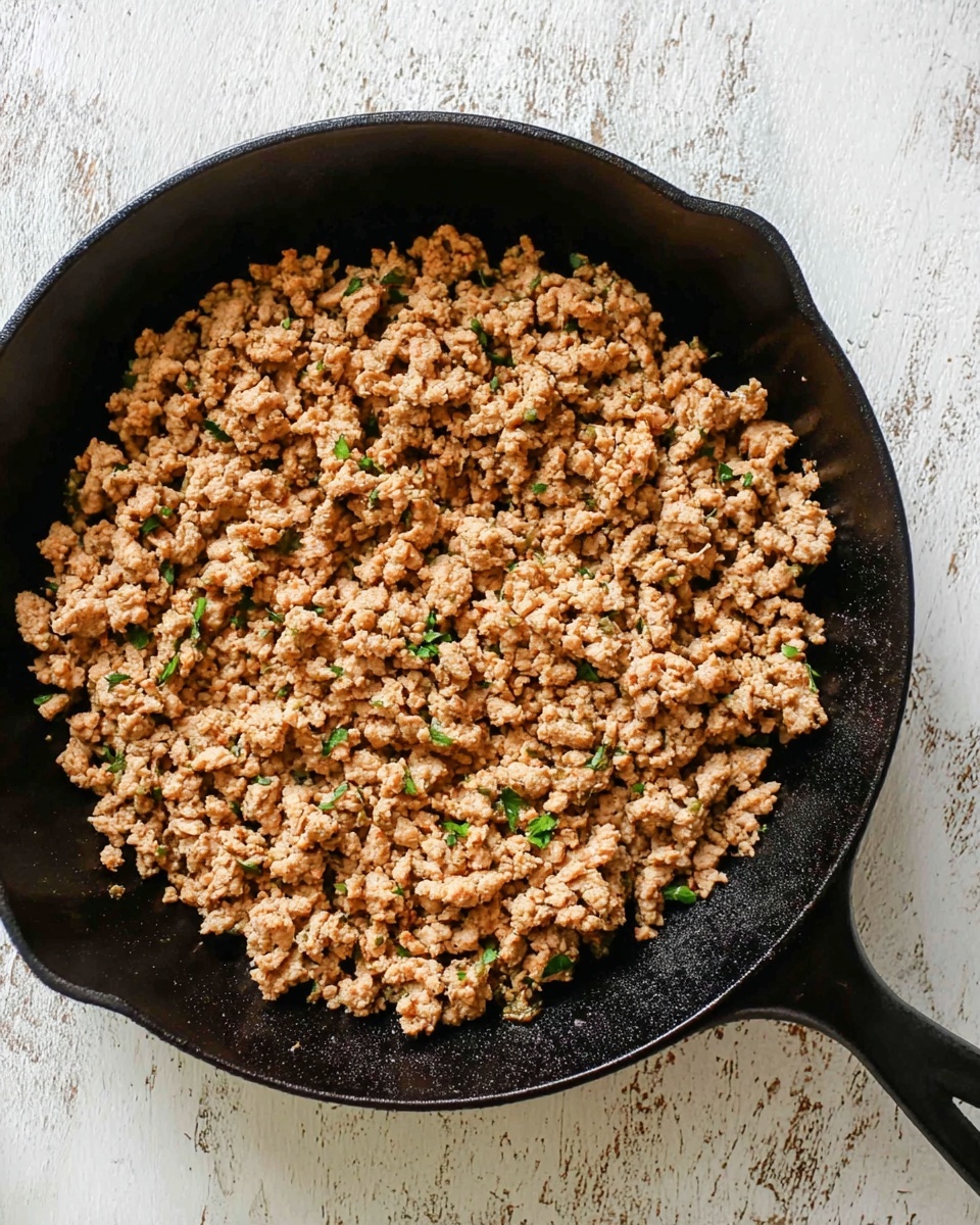 A black cast iron pan filled with cooked ground meat that is light brown with some small green herb bits mixed in. The texture looks crumbly and slightly soft, spread evenly across the pan. The pan is on a white marbled surface with a rustic look. photo taken with an iphone --ar 4:5 --v 7