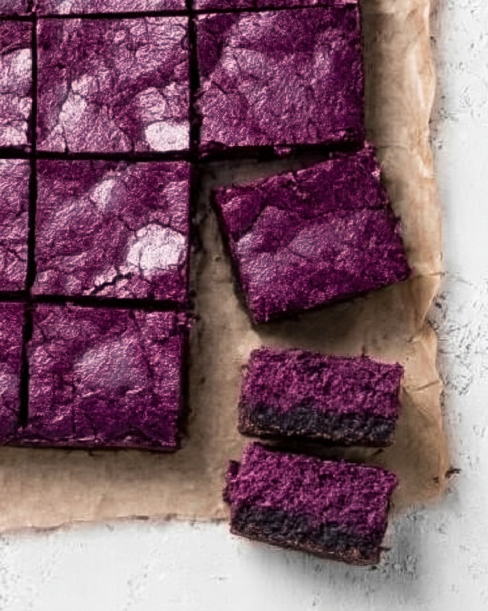 The image shows a baked dessert with a shiny, cracked purple top layer, cut into small square pieces. The dessert is placed on parchment paper, which is on a white marbled surface. Two pieces are separated from the main square block, showing the thickness of the dessert. The purple top layer is firm and textured, with a slightly rough pattern from baking. The photo taken with an iphone --ar 4:5 --v 7