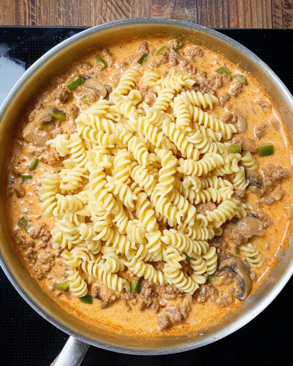 In a stainless steel pan, there is a visible layer of creamy orange sauce mixed with browned ground meat, sliced mushrooms, and pieces of green bell pepper, all cooked together. On top of this sauce layer, there is a thick pile of pale yellow spiral pasta placed in the center, covering most of the sauce beneath. The pan is resting on a black surface with a wooden background visible at the top edge. Photo taken with an iphone --ar 4:5 --v 7