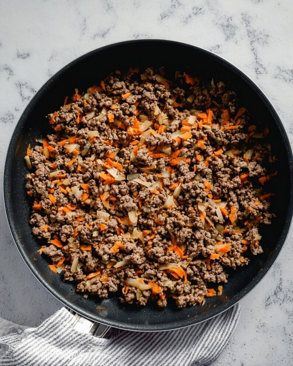 A black frying pan filled with cooked ground meat mixed with small orange carrot pieces and light-colored onion bits, all well combined and evenly spread inside the pan. The pan sits on a white marbled surface with a white and grey striped cloth partially visible at the bottom right. The texture of the meat looks crumbly and tender, with the carrots and onions adding a slight contrast in color and texture, creating a warm and hearty look photo taken with an iphone --ar 4:5 --v 7