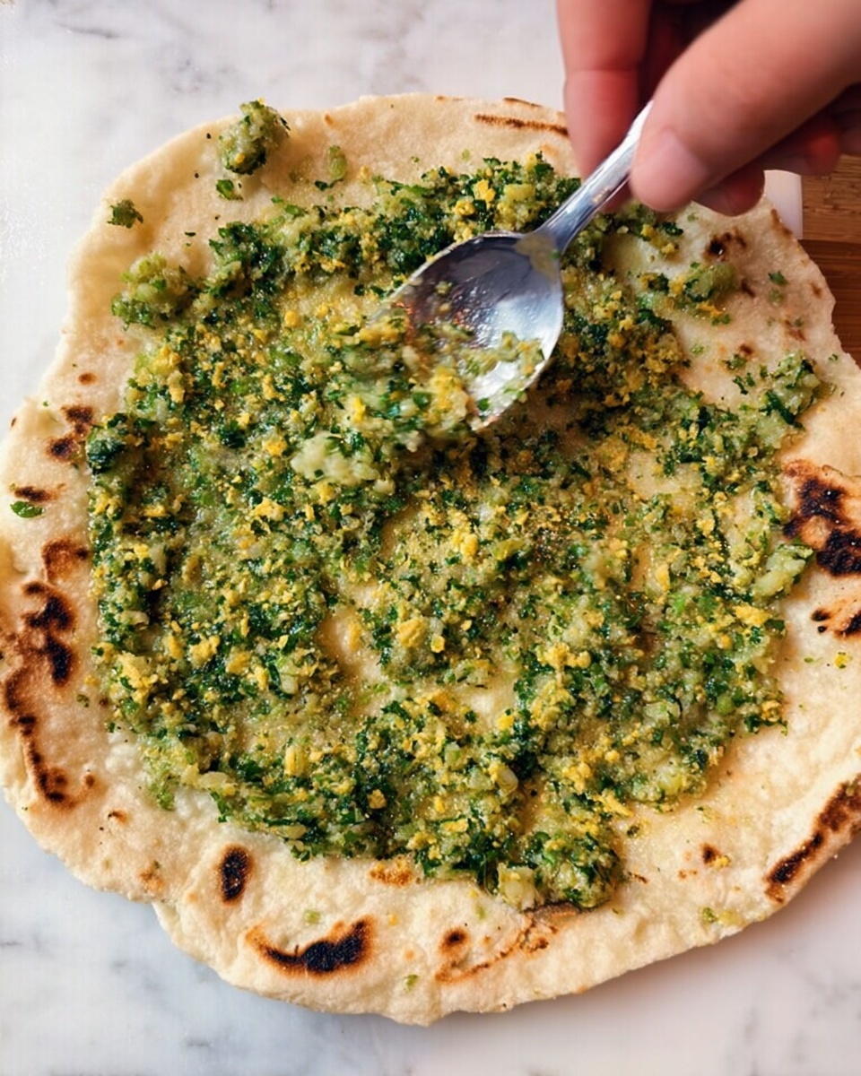 A close-up of a round, light beige flatbread placed on a white marbled surface, with a greenish spread full of small yellow bits evenly covered on top, showing a slightly chunky texture. A woman's hand holds the flatbread steady at the top while another woman's hand uses a shiny silver spoon to spread the mixture. The flatbread has light toasted brown spots around its edges. photo taken with an iphone --ar 4:5 --v 7