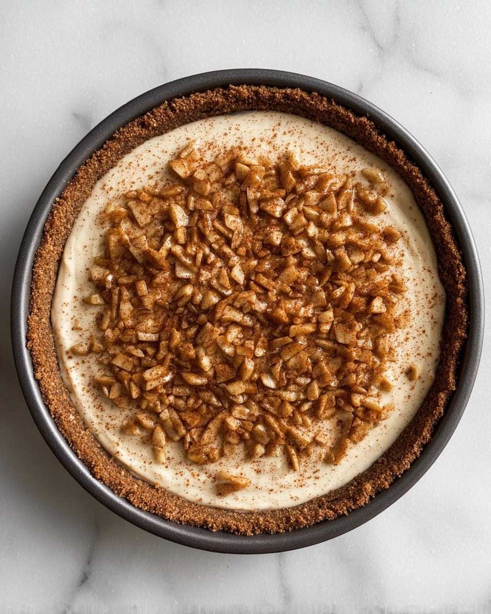 A round pie in a dark pan shows three layers: a thick, crumbly brown crust on the bottom and sides, a smooth, creamy light beige filling in the middle, and a top layer of small chopped apple pieces coated with a dusting of cinnamon, spread evenly. The background is a white marbled surface. photo taken with an iphone --ar 4:5 --v 7
