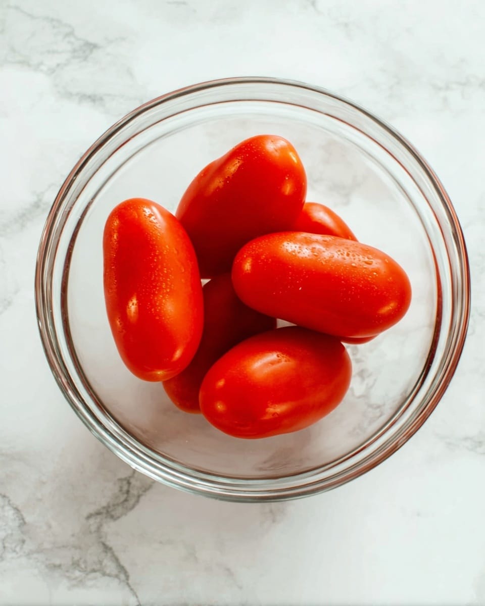 A clear glass bowl sits on a white marbled surface, filled with five bright red, smooth-skinned Roma tomatoes. The tomatoes are plump and arranged close together inside the bowl, showing their shiny texture and slight natural irregularities. The round bowl reflects light softly on its edges, enhancing the vibrant color of the tomatoes photo taken with an iphone --ar 4:5 --v 7