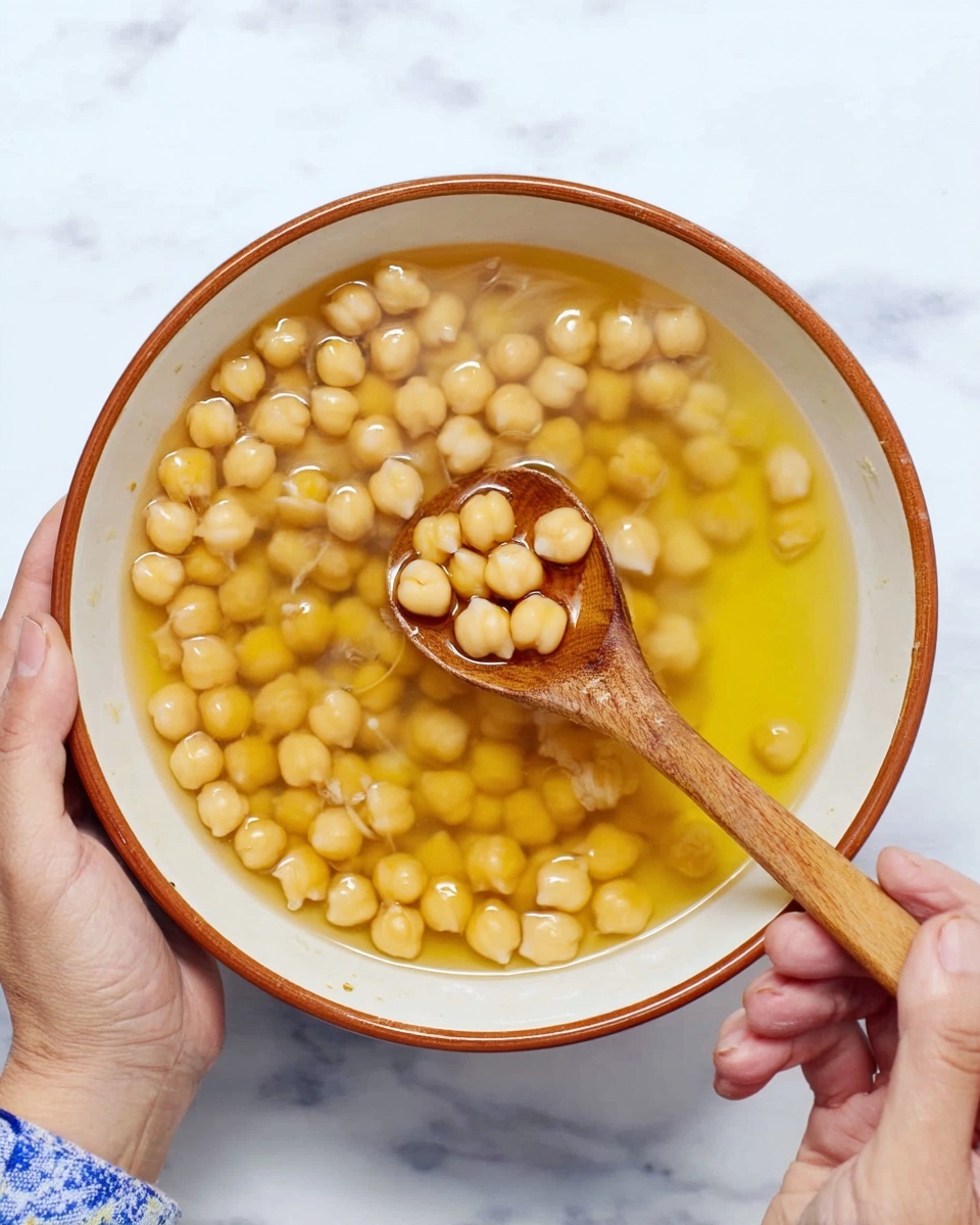 A white bowl filled with round, light beige chickpeas soaking in a clear yellowish liquid, almost filling the bowl. A wooden spoon rests inside the bowl, holding a few chickpeas above the liquid. The bowl is held by both a woman's hand on the left and a woman's hand on the right, against a white marbled surface background. Photo taken with an iphone --ar 4:5 --v 7