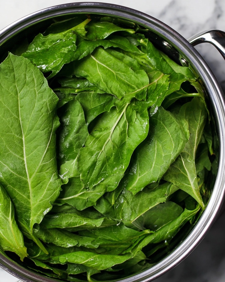 A close-up view of a pot filled with multiple large, fresh green leaves stacked loosely inside. The leaves are shiny with visible veins and a smooth texture, some slightly folded or curved, covering the entire inside surface of the pot. The pot’s shiny silver rim and handles are visible around the edges, resting on a white marbled surface. photo taken with an iphone --ar 4:5 --v 7