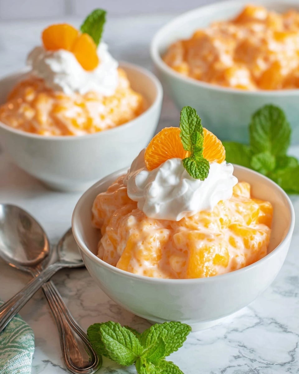 The image shows two white bowls filled with a creamy orange dessert that has a lumpy texture, likely from fruit pieces mixed inside. Each bowl is topped with a dollop of smooth white whipped cream, a bright orange mandarin segment, and a small green mint leaf for decoration. The bowls sit on a white marbled surface with two silver spoons placed beside one bowl. Fresh green mint leaves are also laid next to the closest bowl. In the blurred background, another bowl is partially visible, filled with the same orange dessert. Photo taken with an iphone --ar 4:5 --v 7
