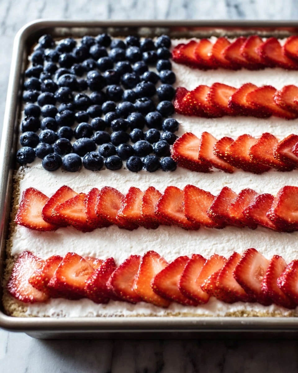 A rectangular dessert in a silver baking tray has three visible layers: the bottom layer is a pale crust, the middle layer is a thick, white cream, and the top layer is decorated like an American flag using fresh fruit. The top left corner is filled with a dense patch of shiny dark blue blueberries, and the rest of the top surface has neat rows of bright red strawberry slices arranged in four horizontal stripes on the white cream. The background surface is a white marbled texture photo taken with an iphone --ar 4:5 --v 7