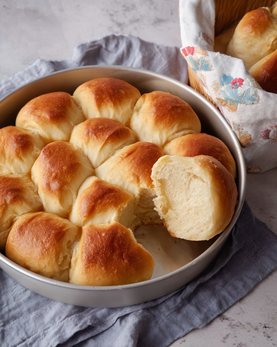 A round white metal pan holds a flower-shaped arrangement of soft, golden-brown baked rolls with a light crust. The rolls are evenly browned on top, showing a smooth, slightly shiny texture. One roll is pulled away and resting on the side, revealing a fluffy, light inside with a moist crumb texture. The pan sits on a light gray cloth, and a white cloth-lined basket with more bread is partially visible in the upper right corner. The surface underneath is a white marbled texture. Photo taken with an iphone --ar 4:5 --v 7