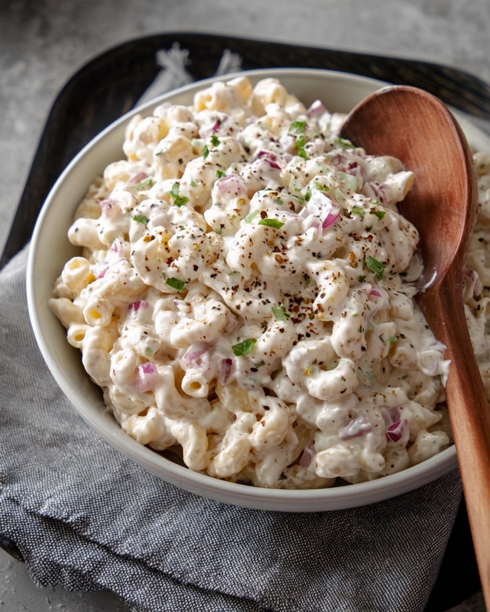A close-up view of a bowl filled with creamy macaroni salad, showing two layers: the bottom layer is white macaroni pasta with some pieces of diced red onion, and the top layer is thick, white creamy dressing mixed evenly with small green herb pieces sprinkled on top, along with some black pepper flakes. The bowl is white and rests on a gray cloth with some black tray edges visible in the background, while a wooden spoon is placed inside the bowl on the right side. Photo taken with an iphone --ar 4:5 --v 7