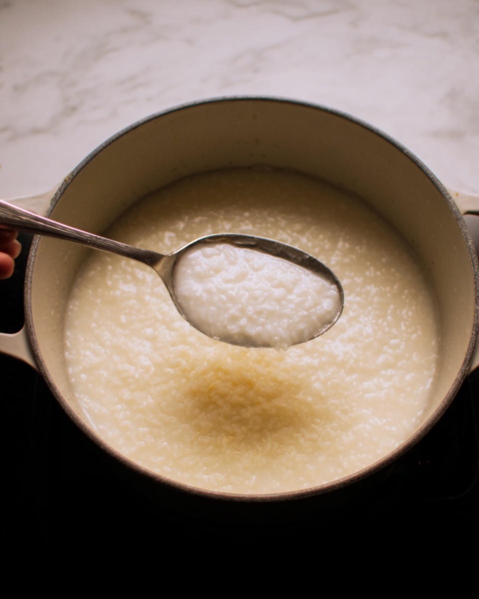 The image shows a white pot filled with soft, thick rice porridge that looks creamy and smooth. A spoon is scooping some of the porridge, showing its slightly grainy texture with small rice pieces spread evenly in the pot. The pot sits on a white marbled surface, and the soft light highlights the creamy white color of the porridge. The silver spoon has a simple design and is held by a woman's hand on the left side of the image. Photo taken with an iphone --ar 4:5 --v 7