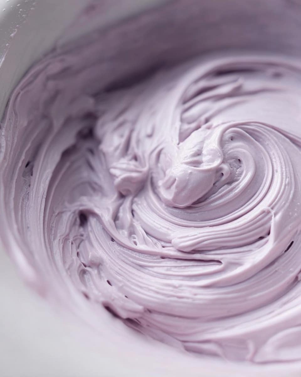 A close-up view of smooth, light purple cream swirled thickly inside a white bowl with a white marbled background. The cream has soft ridges and peaks showing a shiny, whipped texture, gently rolled in circular motions. Photo taken with an iphone --ar 4:5 --v 7