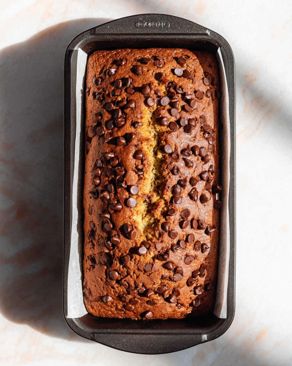 A rectangular chocolate chip banana bread loaf sits inside a black metal baking pan lined with white parchment paper on a white marbled surface. The bread has one main layer that is golden brown and topped with a thick, darker brown crust densely covered in semi-melted chocolate chips. There is a slight crack down the middle of the loaf where the inner yellowish bread is visible. The sunlight casts a sharp shadow of the loaf and pan to the left side. Photo taken with an iphone --ar 4:5 --v 7