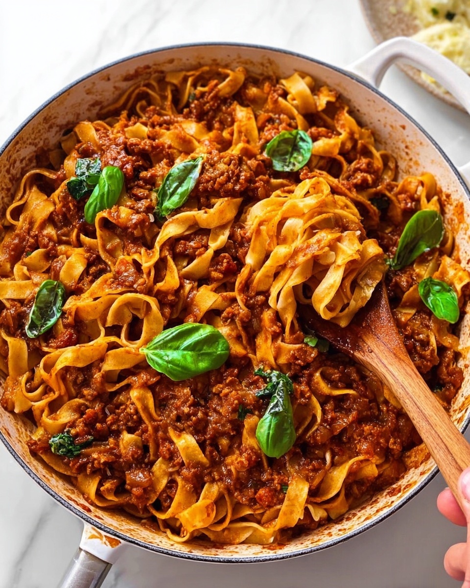 A white pan filled with twisted layers of flat pasta tangled with chunky dark reddish-brown sauce, containing visible small pieces of ingredients mixed in. The sauce covers the pasta unevenly, showing the pasta's smooth, slightly shiny texture. Fresh, bright green basil leaves are scattered on top, adding a burst of color. A woman's hand holding a wooden spoon lifts a twist of pasta from the pan. The scene rests on a white marbled surface. photo taken with an iphone --ar 4:5 --v 7