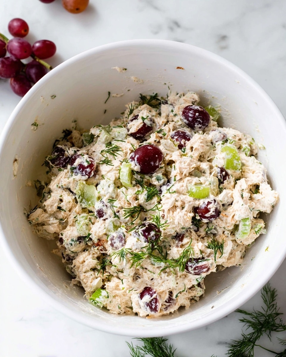 Inside a white bowl, there is a creamy, chunky mixture with layers of light beige chicken salad dotted with small pieces of green celery and red onion. Scattered throughout the salad are whole dark red grapes and bits of fresh green herbs, possibly dill, adding small bright green details. The texture looks thick and mixed but still shows the different elements clearly. The bowl sits on a white marbled surface with a few loose grapes and herb sprigs nearby. photo taken with an iphone --ar 4:5 --v 7