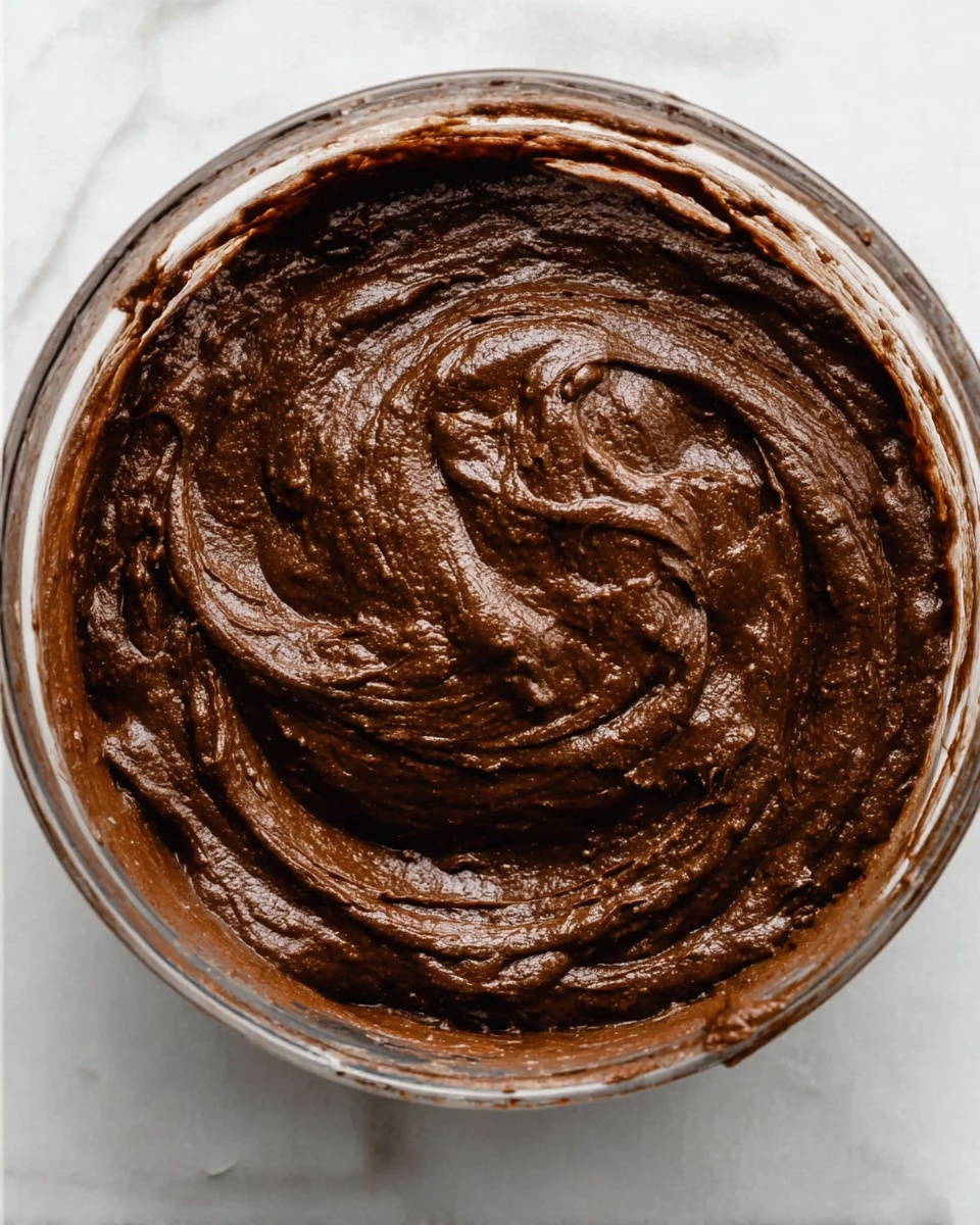 A close-up view of a clear glass bowl filled with thick, dark brown chocolate batter with a smooth, creamy texture. The batter has swirled patterns on its surface, showing a rich and glossy look. The bowl is placed on a white marbled surface, and bits of chocolate batter stick slightly to the sides of the bowl, emphasizing its dense, smooth consistency. photo taken with an iphone --ar 4:5 --v 7
