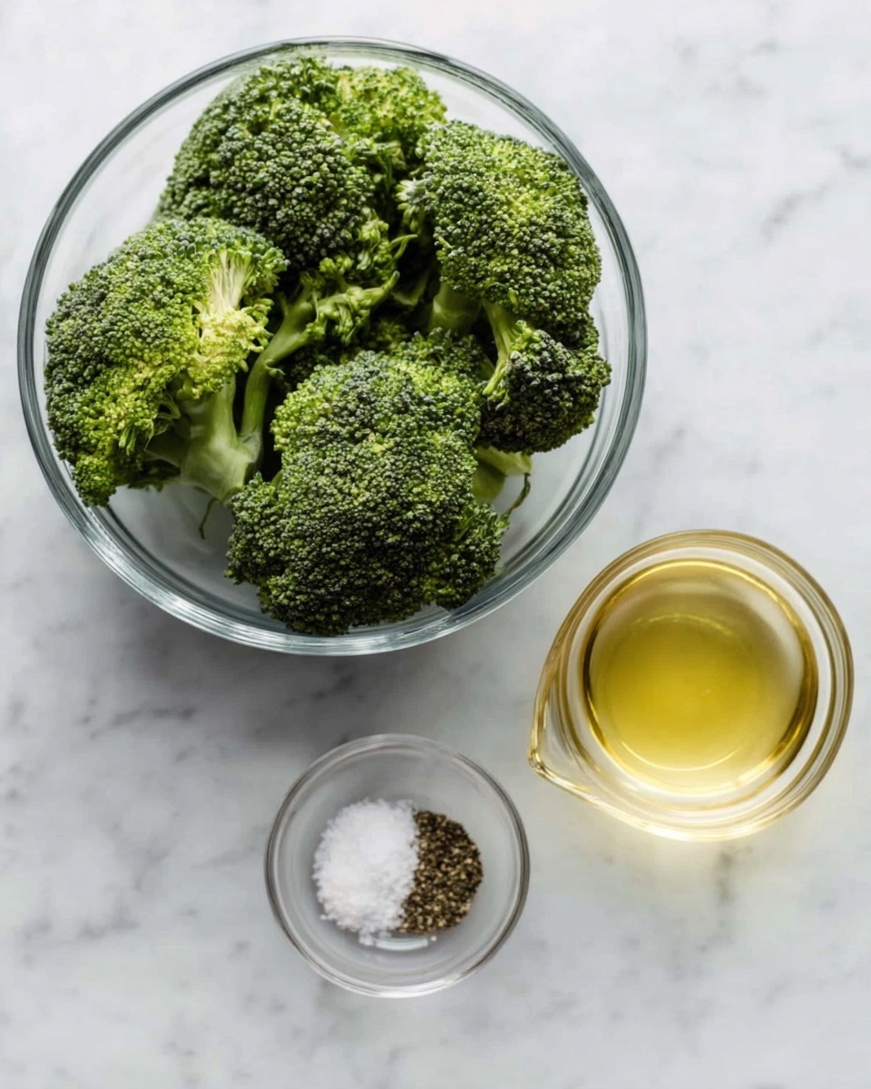 A clear glass bowl filled with several large pieces of fresh, dark green broccoli florets sits on a white marbled surface. To the right of the bowl, there is a small glass cup filled with a light yellow liquid, likely oil or dressing. Below these, a tiny clear bowl holds a mix of white salt and dark pepper. The arrangement of the items is simple and clean, with soft natural light highlighting their textures and colors. Photo taken with an iphone --ar 4:5 --v 7