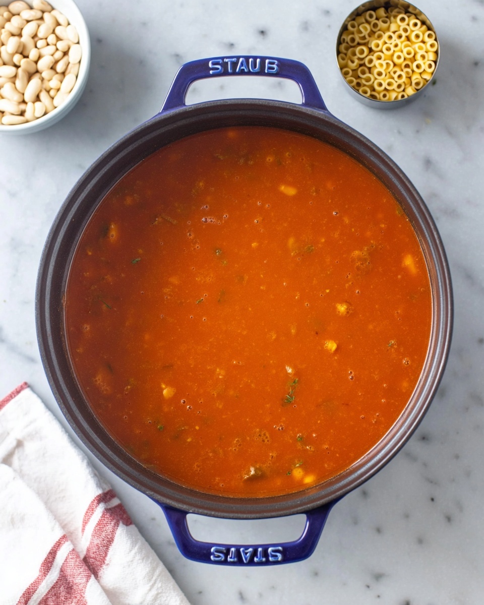 A top-down view of a large deep pot filled with a smooth, orange-red tomato-based soup with small bits of herbs and vegetables visible throughout, sitting on a white marbled surface. Surrounding the pot, there is a white bowl with white beans on the top left and a small metal cup filled with tiny round pasta on the top right. A white cloth with a red stripe is placed on the bottom left corner near the pot. The pot has blue handles with the word
